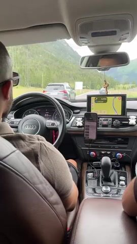 The video shows the interior of a brown-leather-seated car from the back seat, looking forward. A man in sunglasses and a polo shirt is driving an Audi, with his hands on the steering wheel, which has a rosary bead decoration. A navigation screen is visible above the dashboard, and a phone mounted on a holder displays a playlist. The road ahead is winding through a mountainous, forested landscape, with other cars visible in the distance. The car is moving at 88 km/h.