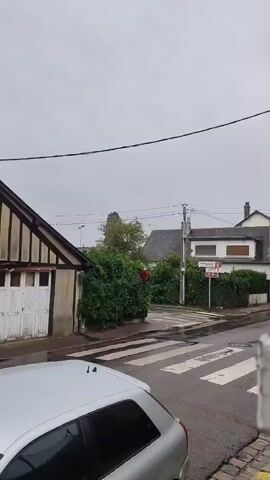 A video pans across a street scene on a cloudy day, likely in a European village. The architecture is distinctively half-timbered, with exposed dark wooden beams against lighter plaster walls. Several houses are visible, some with steeply pitched dark roofs and dormer windows. A silver car is parked in the foreground, partially obstructing the view. The street appears wet, suggesting recent rain, and a pedestrian crossing is marked by white stripes. There are trees and hedges, and utility poles with wires are visible against the overcast sky. A red traffic sign and a white sign with red lettering are mounted on a pole at an intersection. One house has a long, dark and white banner hanging from an upper window. No people are clearly visible in the video.