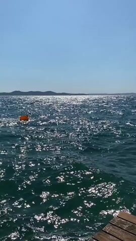 A still shot of the ocean with waves and a wooden pier. There are distant buildings and a clear blue sky.