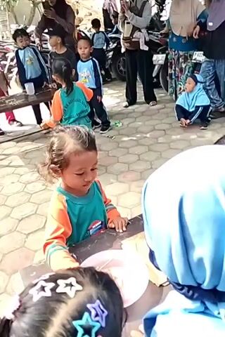 In a daylight outdoor scene, a group of young children and adults are gathered around a table with a tiled ground. A young girl in an orange and teal shirt is focused on something held between her fingers, possibly a craft or game. To her right, a child wearing a blue hijab is facing away from the camera. Behind them, other children in similar attire are visible, some standing and others sitting. Adults are scattered throughout the background, with some holding phones and engaging with the children. Several motorcycles are parked behind the group, suggesting a community event or gathering. The overall atmosphere appears to be one of activity and engagement, with a focus on the children's participation.