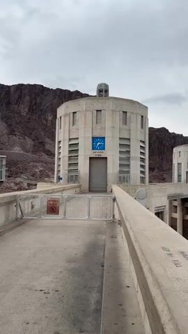 The video shows a view of the Hoover Dam. In the foreground, there is a concrete walkway with a railing and a metal gate with a "Do Not Enter" sign. Behind the gate is a large, cylindrical concrete structure with a clock on it that reads "NEVADA TIME." The structure has several horizontal windows that appear to be louvers. The background features a rocky mountain landscape and a body of water, likely Lake Mead. There are several cars and people visible in the distance. The sky is overcast. The camera pans slightly to show more of the dam and the surrounding environment.