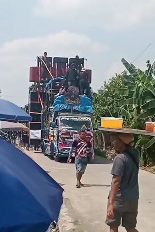 A man walks towards the camera carrying a tray of drinks on his head. Behind him, a large truck is covered in speakers and decorated with blue fabric. Several people are standing on top of the speakers. Another man walks behind the first man, looking towards the camera. A blue tent is visible to the left, and trees line the right side of the road. The scene appears to be outdoors on a sunny day.