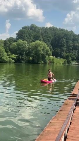 A man is paddleboarding on a calm lake. He is shirtless and wearing black shorts. He is holding a paddle and appears to be propelling himself forward. The paddleboard is pink and yellow. In the background, there is a lush green forest and a blue sky with white clouds. The sun appears to be shining, indicating it is daytime. A wooden dock is visible in the foreground, partially obscuring the view of the lake. The water is a murky green color with some ripples. The man seems relaxed and is enjoying the activity. The scene is tranquil and natural, suggesting a summer day in a rural or park-like setting.
