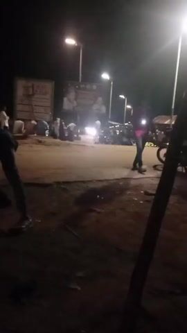 The video shows a dimly lit outdoor scene at night. Several streetlights illuminate a dirt or unpaved road. There are people present, some gathered in small groups. Several motorcycles are visible, parked or being ridden. There are large posters or banners displayed on structures in the background. The overall impression is that of a public gathering or market area at night.