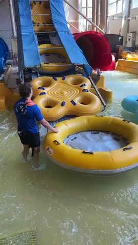 A lifeguard wearing a blue shirt and black shorts stands in shallow water in a water park, holding a yellow inner tube. Behind him, a large blue water slide descends into the water, with yellow inner tubes stacked on its side. To the left, a person rides down a blue tube slide, splashing into the pool. To the right, a red tube slide curves into view, and other yellow inner tubes are visible. Another person, wearing a blue swimsuit, walks through the water with a light blue inner tube. The water is murky green, and the environment appears to be indoors with bright lighting and structural elements of a water park visible.