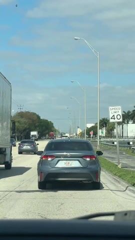 The video depicts a car driving down a road, with a gray Toyota Corolla in front. The Toyota Corolla has a Florida license plate that reads "34E JZI." The speed limit is 40 miles per hour. There are streetlights along the side of the road, as well as some palm trees and other greenery. There is a large truck on the left of the vehicle that recorded the video. There is a crosswalk sign. The sky is partly cloudy. There are other cars in the distance, and traffic appears to be moving at a steady pace.