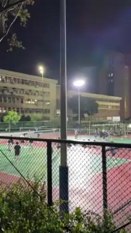 At night, behind a chain-link fence, several illuminated outdoor sport courts are in use. Groups of people are engaged in sporting activities, possibly volleyball or badminton. Buildings stand in the background under a dark sky. The camera pans from one court to the next, revealing the extent of the facility and the activities underway. Foliage in the foreground partially obscures the view at times. Electrical poles and wires are also visible in the foreground.