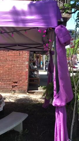 A medium shot shows an outdoor market stall adorned with purple fabric and string lights. A brick wall forms the backdrop. To the left, a round table is covered with a clear plastic cloth featuring a floral design. In the center, a large tapestry or fabric hangs, depicting a young woman with a flower crown and large blue eyes, posed against a colorful, abstract background. To the right of the tapestry, a birdcage filled with greenery hangs from the tent's ceiling. A small table to the right displays a few items and pink decorative elements. The scene is bathed in sunlight, creating shadows from the tent and foliage.