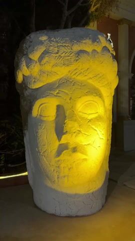 The video shows a large, white, heavily textured sculpture of a human head, illuminated by a strong yellow light. The camera zooms in on the face, highlighting the details of the rough surface and the shadows cast by the light. The sculpture is outdoors, possibly in a courtyard or garden. Behind the sculpture, there is some greenery and a building with a red facade and columns. The overall scene is somewhat abstract due to the close-up perspective and the stark lighting.