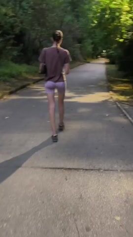 A person is running on a paved path through a wooded area. The person is wearing a purple t-shirt and light purple shorts. The person is casting a shadow on the path. The sun is shining through the trees, creating dappled light on the path. The person is running away from the camera.