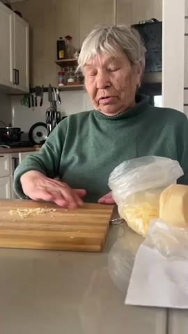 An elderly woman with short gray hair stands in a kitchen, looking directly at the camera and talking. She is wearing a green turtleneck sweater. In front of her on a beige countertop is a wooden cutting board with some small bits of food on it. Next to the cutting board are plastic bags containing shredded cheese and a chunk of cheese. The kitchen has white cabinets and beige tile walls. A shelf above the counter holds bottles and jars. A rack of knives hangs on the wall. A pot sits on the stove in the background. The woman gestures slightly with her left hand as she speaks.