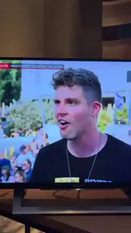The video shows a man being interviewed on television. The interview is taking place live from Hostages Square in Tel Aviv. The man is wearing a black t-shirt with white text on it and a silver necklace. He has short, curly hair. In the background, there is a large crowd of people holding flags. The interviewer is a Black man wearing a dark blue shirt. He is holding a piece of paper and a pen. The interview seems to be taking place outdoors in a public space. The TV on which the interview is being shown is resting on a wooden stand.