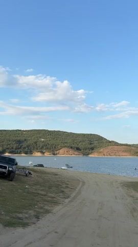 The video begins with a view of a dirt road leading down to a body of water. The water is calm and reflects the clear blue sky with scattered white clouds. On the left side of the frame, there is a patch of dry grass and dirt, with a few objects that could be vehicles or boats parked near the water's edge. The background is dominated by rolling hills covered in green trees and vegetation.

As the video progresses, the camera pans to the right, revealing more of the shoreline and the water. Several boats are visible, some docked and others floating on the water. A car is parked on the grassy area near the water. A "no swimming" sign is visible in the middle of the frame. In the distance, more hills can be seen.

The camera continues to pan right, showing a wider expanse of the lake and the surrounding landscape. More boats are scattered on the water, and a larger structure that looks like a floating platform or a house on water is visible. A white car is parked near this structure. The hills in the background appear to be a mix of green vegetation and areas with exposed earth. The sky remains clear blue with some wispy clouds. The video ends with a panoramic view of the lake, the shoreline with parked vehicles and boats, and the tree-covered hills under a bright blue sky.