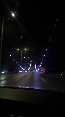 The video shows a view from inside a vehicle driving at night on a highway towards a suspension bridge illuminated with lights. The bridge's towers are red and white, and the cables are strung with white lights. Streetlights line the highway, and there are other vehicles on the road, including a truck with its hazard lights flashing. The camera lens appears to have some smudges or dirt on it, creating small flares and distortions around the light sources. The overall impression is of a nighttime journey across a brightly lit bridge.