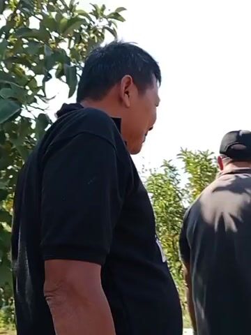 The video begins with a close-up of the back of a man's head and shoulders as he walks away from the camera. He is wearing a black t-shirt and tan pants. The camera then pans to the right, revealing another person walking ahead of him, also from behind. This person is wearing a dark gray t-shirt and shorts, with a black baseball cap. They are walking on a dirt path lined with vegetation. In the background, there are trees, some construction materials including rebar structures, and a clear sky. The camera continues to follow the two individuals as they walk further into the scene. Additional people, partially obscured, can be seen walking behind the first two. The overall environment appears to be outdoors in a rural or semi-rural setting, possibly under construction.