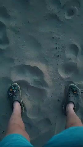 The video opens with a close-up shot of a person's legs wearing blue shorts and grey Crocs adorned with colorful charms, standing on a sandy surface with footprints. The camera then pans up to reveal a blurry beach scene with the ocean and a treeline in the distance, illuminated by a colorful sunset. The sky transitions from a deep blue to orange and yellow hues.