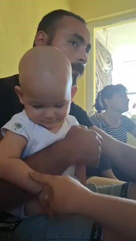 This close-up video, filmed in Konevets, Bulgaria, captures a tender moment between a baby and an adult. The baby, with a shaved head and wearing a white onesie with a small palm tree emblem, is being held by an adult male, who is partially visible in the frame. The adult's arm encircles the baby, providing a sense of security. Another adult, a woman, is visible in the background, turned away from the camera and looking towards a window or bright light. The scene appears to be indoors, with a yellow wall visible behind the adult male. The lighting suggests it is daytime. The baby's gaze is directed downwards, towards the adult's hand, which is gently touching the baby's arm. The overall mood is calm and affectionate. No visible text is present in the video.