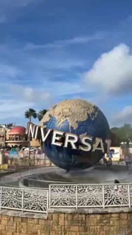 The video shows the Universal Studios globe logo in Orlando, Florida. The globe is blue and tan, with the word "UNIVERSAL" wrapped around it. The globe is surrounded by a fountain and a white fence. In the background, there are buildings and palm trees. The sky is blue with white clouds. There are people walking around in the background.
