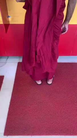 A person wearing a maroon robe and sandals stands in front of a yellow and red wall with a red mat on the floor. The person is facing away from the camera.