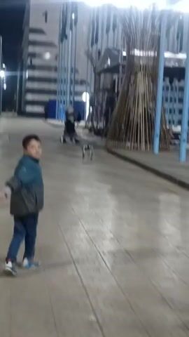 The video opens with a young boy wearing a blue jacket and jeans looking at the camera, then pans to the right, revealing an open paved area at night. There is a small dog walking in the distance, toward a structure made of bamboo poles and light blue metal supports. People can be seen walking around the area. In the background, there's a building with the number "1" on it and several flagpoles. A man in a black suit walks in the opposite direction of the dog.