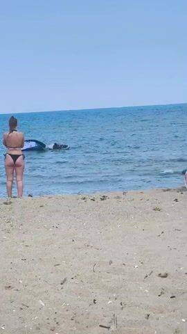 A woman in a black bikini stands on a sandy beach, facing a calm blue ocean. A jet ski is partially submerged in the water to her left, with a person lying on it. The sky is clear and blue. The video is short, with the scene remaining mostly static.