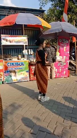 A video clip shows a woman in a traditional patterned dress and sneakers dancing in front of food stalls. She is facing the camera and appears to be posing. Behind her, a young girl in a pink shirt walks by, followed by a woman in a hijab. In the background, there are several food stalls with colorful umbrellas and signage. One stall advertises "Es Teler" (shaved ice dessert), and another has a sign for "Cikibul" (a type of snack). A man in a plaid shirt is visible behind the dancing woman, and another man in a red shirt is walking to the left of the frame. The ground is paved with brick-like stones. The lighting suggests it is daytime.