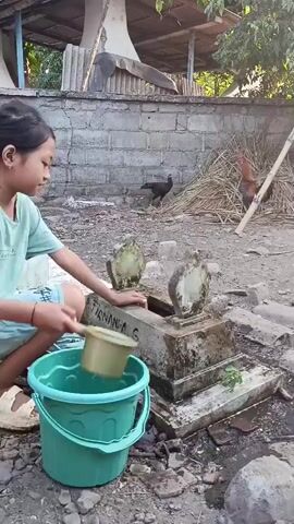 A young girl kneels by a grave, pouring water from a small cup onto it. She is wearing a light blue t-shirt and light blue shorts, with her hair tied up. A green bucket sits on the ground next to her. The grave is made of concrete and has inscriptions on it. Several chickens are visible in the background, as well as a pile of hay and some trees. The scene appears to be outdoors in a rural or semi-rural setting.