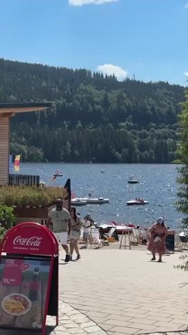 The video shows a waterfront scene on a sunny day. In the foreground, a red Coca-Cola advertisement is partially visible. Several people are walking along a paved walkway next to a body of water. On the left, a man and a woman are walking away from the camera, with the man wearing a striped shirt and shorts, and the woman in a floral dress. Further back, other people are seen near beach chairs and umbrellas. The lake itself is calm with several small boats and watercraft on it, including what appear to be paddle boats. In the background, a lush, tree-covered mountain slopes down to the water. The sky is blue with a few scattered clouds.