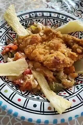 A close-up shot shows a plate of food with a breaded chicken cutlet on top of a salad. The salad appears to be made with chopped tomatoes, bell peppers, and possibly onions, and is topped with what look like potato wedges. The plate has a black and white geometric pattern with red and blue dots around the rim. Another plate with breaded chicken is visible on the right side of the frame, resting on a white paper napkin. The food is placed on a table with a patterned tablecloth. The lighting suggests it could be daytime. No people are visible in the frame.