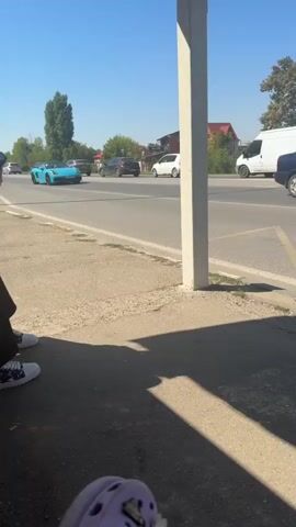 A turquoise convertible sports car with two people inside drives past a person sitting on a curb. The car is on a road with other vehicles, and there are buildings and trees in the background. The sky is blue and clear. The person on the curb is wearing white sneakers with black and white laces and is partially visible in the bottom left corner of the frame. A purple object, possibly a shoe or sandal, is visible in the very bottom center of the frame. The scene appears to be outdoors on a sunny day.