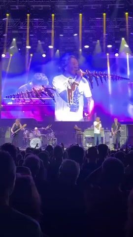 The video shows a live concert performance by the band Deep Purple. The band members are on stage, with a large screen behind them displaying footage of the drummer. The audience is visible in the foreground, cheering and raising their hands. The stage is lit with bright purple and blue lights, and there are several spotlights shining down. The drummer is shown playing his kit vigorously. The lead singer is in the center of the stage, holding a microphone and singing. The other band members are playing their respective instruments, including guitar and keyboards. The overall atmosphere appears energetic and enthusiastic.