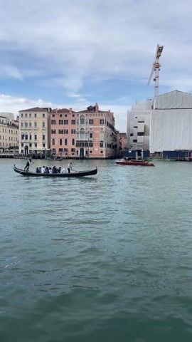 The camera captures a daytime scene in Venice, Italy, showcasing a gondola carrying a group of people down a canal. The gondola is manned by two gondoliers, one at the front and one at the back, both dressed in white shirts. The passengers are mostly seated and appear to be enjoying the ride.

The backdrop includes buildings of various colors and architectural styles, lining the canal. To the right, a building is covered in white tarp, with a construction crane visible behind it. Another smaller, wooden motorboat is also on the canal.

The water in the canal is a greenish-blue color, with slight ripples on the surface. The sky is partly cloudy, with a mix of blue and white.