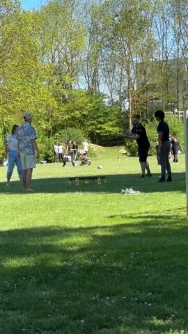 This video captures a vibrant scene of people playing Spikeball on a sunny day in a grassy park in Ditzingen, Germany. In the foreground, four young adults are actively engaged in the game around a black Spikeball net. A man in a patterned shirt and shorts is positioned near the net, while another man in a dark shirt serves the small yellow ball, hitting it off the net. A woman in a light top and blue jeans, and another person in a dark outfit, are also part of the play, observing the serve and preparing to react. Their casual attire and bare feet or athletic shoes suggest a relaxed, recreational atmosphere.

The setting is an open green space, indicative of spring or early summer, with numerous trees displaying lush green foliage in the background under a clear sky. Strong shadows cast on the grass indicate bright sunlight. In the mid-ground and background, other individuals are visible, some sitting on the grass, others standing, suggesting a larger gathering or general park activity. A building can be partially seen in the far background on the right. The overall mood appears cheerful and active, with participants enjoying the outdoor sport. No visible text is present in the video.