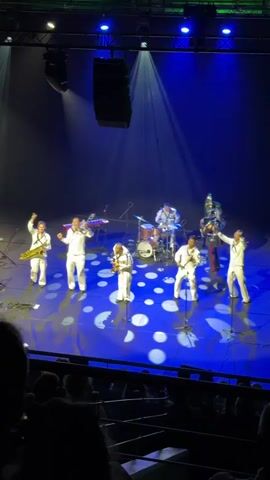 A full shot captures a band performing on a stage in front of an audience. The stage is illuminated with blue and green spotlights, creating a dramatic effect. The band members, dressed in matching white uniforms, are positioned across the stage. Several musicians play brass instruments, including saxophones and a tuba. One member is behind a drum set. The stage floor has a pattern of white circles. The audience is visible in the foreground, seated in dark seats, with only the tops of their heads and shoulders visible. The band members appear to be actively engaged in their performance, with some raising their instruments and arms.