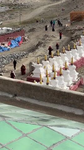 The video shows a scene with multiple white, stupa-like structures with gold tops arranged in rows. Several people, including some wearing maroon robes, are walking on a dirt path in the background. Some of the path leads uphill. There is a sparse, rocky landscape with some remaining snow in the background. A blue tarp covers materials to the left, and a wooden structure is on the right. The camera is positioned above the scene, looking down at the stupas and the path.
