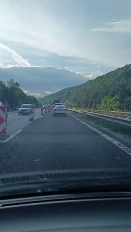 The video shows a first-person perspective of a vehicle traveling on a highway. The road appears to be undergoing some sort of construction or maintenance, as evidenced by the presence of traffic cones and barriers lining one side of the highway. Several cars are visible on the road ahead, all moving in the same direction. The sky above is partly cloudy, with the sun shining through in some areas. The surrounding environment is hilly and covered with trees. The camera is positioned at dashboard level, looking forward through the windshield.