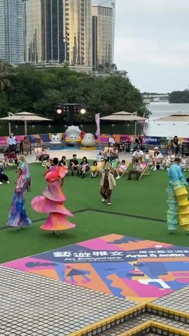 The video shows a performance taking place outdoors in front of an audience. Three performers are present. Two are on stilts, wearing colorful, elaborate costumes. One is in the center, wearing a brown suit and hat, and appears to be the emcee. The audience is seated in chairs and on the ground, facing the performance area. Behind the audience, there is a stage setup with cartoon characters and lighting. In the background, there are tall buildings, trees, and a body of water. The performance area is covered with artificial grass and a large, colorful square design with text in both Chinese and English that says, "Art Everywhere, A Cultural Vibe at the Weekend." The sky appears overcast.