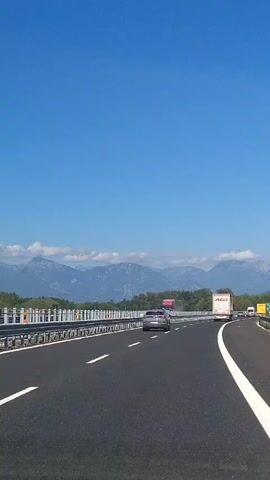The video shows a view from inside a vehicle traveling on a highway. The highway has multiple lanes, marked with white lines. There are other vehicles on the road, including cars and trucks. On the left side of the highway, there is a metal guardrail. In the distance, there are mountains and trees. The sky is clear and blue. The sun appears to be shining. The video is taken from a slightly elevated perspective, looking forward along the road.