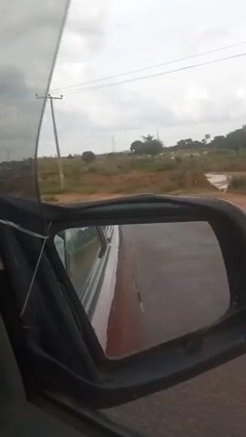 The video shows a view from the inside of a car, looking out through the side mirror and the open door. The car appears to be moving on a paved road, with a rural landscape visible outside. There are trees, including palm trees, and some power lines in the background. The sky is overcast. The side mirror reflects the road behind the car, and the side of the car itself. The focus is on the movement and the scenery passing by.