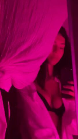 The media file is a video of a young woman taking a selfie in a mirror. She is wearing a white shirt over a black bra. The lighting is a bright pink hue, creating a dramatic and intimate atmosphere. A sheer, white curtain is draped to the left of the frame, partially obscuring the woman and adding a soft, diffused texture to the image. She is holding a smartphone in her right hand, angled to capture her reflection. Her expression is calm, with her eyes closed or looking down. The background is dark, with some faint hints of a room. The overall mood is sensual and artistic, likely captured indoors at night. There is no visible text in the media file.