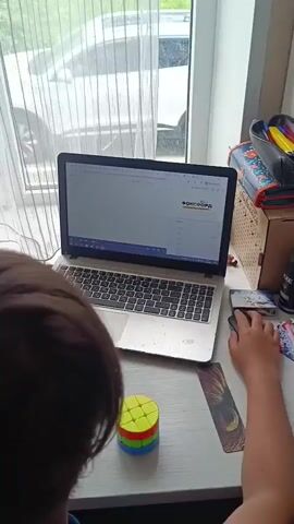 A young person is sitting at a white desk facing a laptop. The laptop is open and displaying a webpage with the word "ФОКСФОРД" visible in the upper right corner. A hand is on the right side of the laptop, holding and using a computer mouse. There is a cylindrical Rubik's cube on the desk to the left of the laptop. A smartphone is to the left of the laptop, and a window with a white sheer curtain is behind the laptop. Outside the window, greenery and a blurred image of a car can be seen. To the right of the laptop are school supplies in a wooden box. The young person is wearing a blue shirt. The desk has a bookmark and some other small items scattered around it.