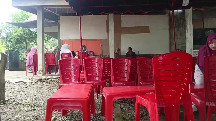 The video shows a collection of red plastic chairs and tables arranged outdoors. A woman wearing a purple hijab is visible on the right side of the frame, interacting with the chairs. In the background, several other people are seated on red chairs, some of whom are also wearing hijabs. The environment appears to be a covered outdoor area, possibly a gathering space or a waiting area, with some trees and buildings visible in the distance. The ground is covered with gravel and dirt. The lighting suggests it is daytime.
