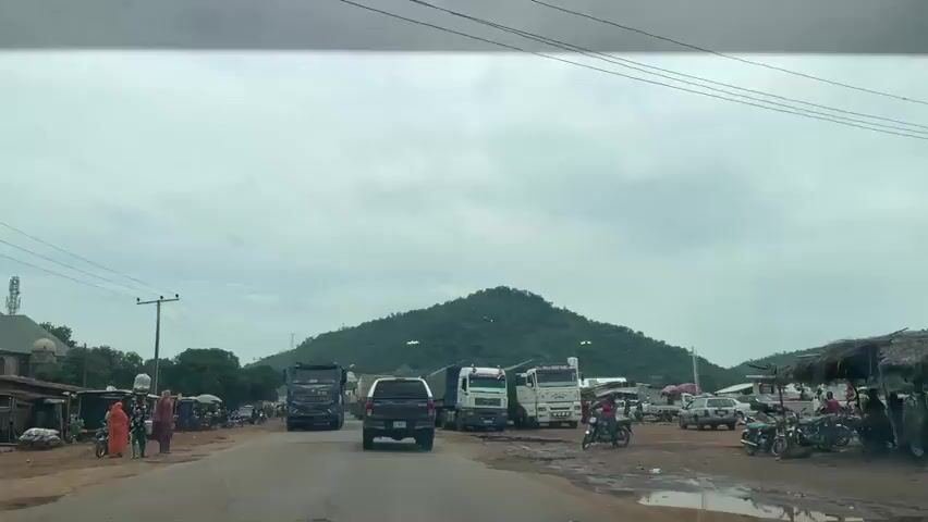 The video begins with a wide shot of a road with several large trucks parked or driving on it. In the background, a large, green, tree-covered hill dominates the landscape under a cloudy sky. On the left side of the road, there are some small buildings and a few people walking. As the video progresses, the perspective shifts, showing more of the trucks and the surrounding area. Several of the trucks are white, while one in the foreground is dark blue. There are also cars and motorcycles visible. Some people are standing near the trucks, and a few individuals are riding motorcycles. The road appears to be unpaved in some sections, with puddles of water visible. On the right side of the road, there are some makeshift stalls or shelters. In the far distance, there are more hills and some structures, possibly a gas station. The overall scene depicts a busy roadside area with a lot of heavy vehicle traffic and some commercial activity.