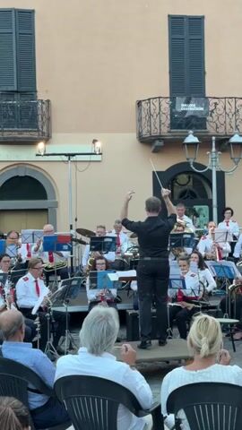 A band plays music outdoors for an audience. A conductor stands on a raised platform and directs the band with a baton. The band members are all wearing matching white shirts and red ties, and some are playing brass instruments, woodwinds, and drums. The audience is seated in chairs in front of the band. The scene is lit by natural light and some artificial lights. A building with balconies is visible in the background.