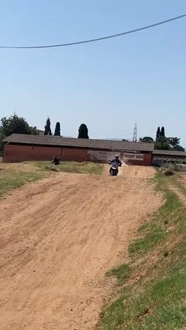 Here is a description of the video.

The video shows a person riding a motorcycle on a dirt track. The rider is wearing a helmet, gloves, riding boots, and riding pants. They jump the motorcycle over a mound of dirt and continue on the track. Three people are watching. The track is surrounded by grass, trees, and buildings. The sky is blue and clear.