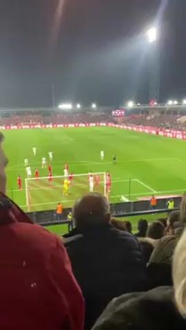 A video taken from stadium seating shows a soccer game in progress at night. The stadium is mostly full with the majority of the crowd out of focus. On the field, players in white uniforms face players in red uniforms on the right side of the field in front of a goal that is closest to the viewer. The goal keeper, dressed in yellow, stands ready. A player in white is standing in front of the goal. There are several people standing and seated in front of the person taking the video, blocking the view of the lower left side of the field.