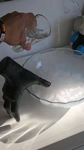 A close-up shot shows a hand wearing a black leather glove, adorned with stitched detailing and a single red fingernail, poised above a clear glass bowl filled with ice. A clear glass of liquid is tilted, pouring a stream of water into the bowl, causing a thick white vapor to emerge and engulf the ice. The background is blurred but suggests a bright, indoor setting with a white surface and a hint of a child's doll with clown-like features. The hand holding the glass of water is also visible, wearing a wristband with colored stripes.
