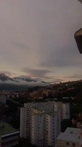 The video pans across a city skyline at dusk, with a cloudy sky and a mountainous backdrop. Several apartment buildings are visible in the foreground and mid-ground, some appearing to be identical in design. The mountains in the background are partially obscured by thick clouds, with hints of pink and orange light suggesting the setting sun. The overall impression is of a sprawling urban landscape transitioning into nighttime.