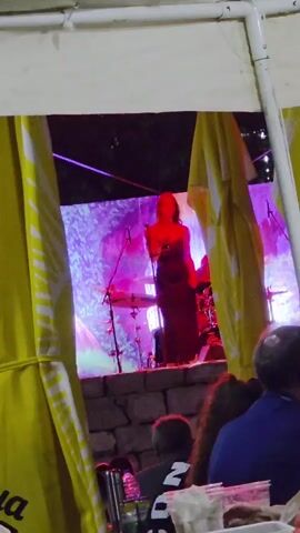 A woman in a long dark dress stands on a stage and sings into a microphone. She is illuminated by purple and pink stage lights against a projected backdrop of abstract colors and shapes. Behind her, a drummer is visible. In the foreground, the backs of several people's heads are visible as they sit at tables. Yellow fabric curtains frame the view of the stage.
