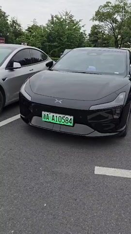 The video shows a close-up view of two parked electric cars, a black Xpeng P7 and a silver Tesla Model 3, from the front. The Xpeng P7 is on the right, and the Tesla Model 3 is on the left. Both cars are parked in designated parking spots marked with white lines on an asphalt surface. In the background, there are trees and other vehicles, including a white SUV and other cars partially visible. The sky is overcast. The license plate of the Xpeng P7 reads "A A10584" and is green. The license plate of the Tesla Model 3 reads "A DQ6187" and is also green. The camera pans slightly to focus on the front of each car.